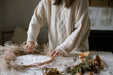 Woman making stylish autumn wreath. Hands with dried grass, herbs, leaves, ribbon and scissors on...