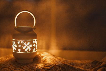 Glowing christmas lantern with golden lights on cozy knitted sweater on frosty window sill in eve....