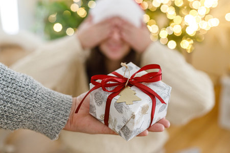 Merry Christmas and Happy Holidays! Hand holding christmas gift box on background of happy woman with closed eyes at christmas tree with golden lights. Exchanging presents surpriseの写真素材