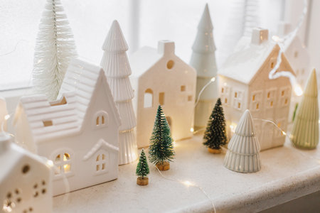 Christmas still life, miniature winter holiday village. Merry Christmas and Happy Holidays! Christmas glowing white little ceramic houses and snowy trees on window. Festive stylish decorationの写真素材