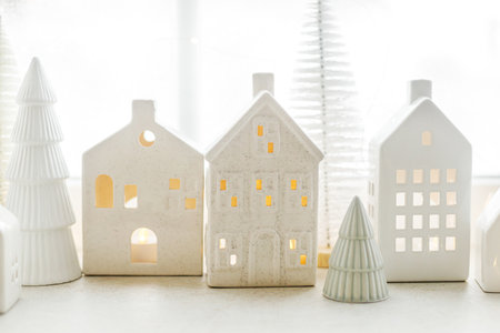 Christmas still life, miniature winter holiday village. Merry Christmas and Happy Holidays! Christmas glowing white little ceramic houses and snowy trees on window. Festive stylish decorationの写真素材