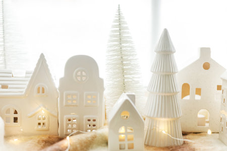 Christmas little ceramic houses, snowy trees and lights on cozy scarf on window. Xmas background. Merry Christmas! Christmas scene, miniature holiday village. Holiday modern decorationsの写真素材