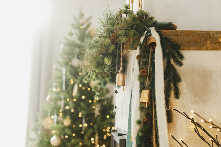 Stylish christmas decorated fireplace mantel with fir branch, bells, ribbon on background of christmas tree with lights in modern living room. Atmospheric holiday at home. Merry Christmas!の写真素材