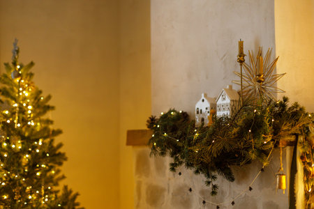 Stylish christmas decorated fireplace mantel with fir branch, bells, ribbon and garland light on background of christmas tree in modern living room. Atmospheric eve at home. Merry Christmasの写真素材