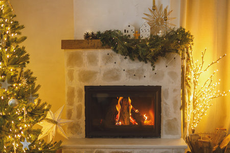 Stylish decorated christmas tree with vintage ornaments and modern decor of fireplace mantel with bells, ribbon and garland lights in evening. Atmospheric christmas eve at home. Merry Christmas!の写真素材