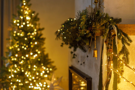 Stylish christmas decorated fireplace mantel with fir branch, bells, ribbon and garland light on background of christmas tree in modern living room. Atmospheric eve at home. Merry Christmasの写真素材