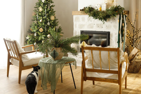 Cute cat sitting in stylish christmas decorated living room with christmas tree and modern fireplace mantel with fir branch, bells and garland light. Atmospheric holidays at home. Merry Christmas!の写真素材