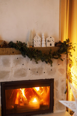 Stylish christmas decor of fireplace mantel with fir branch, ornaments, bells, ribbon and garland lights in evening. Atmospheric christmas eve at home. Merry Christmas!の写真素材
