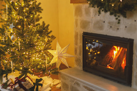 Merry Christmas! Stylish decorated christmas tree with vintage ornaments, garland lights, star, gifts and burning fireplace in evening. Atmospheric christmas eve at homeの写真素材