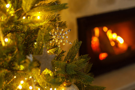 Beautiful decorated christmas tree with glass snowflake, vintage baubles and golden lights on background of burning fireplace. Winter holidays preparation. Atmospheric magical eveの写真素材
