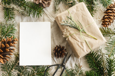Christmas card mock up. Empty greeting card and stylish zero waste gift on rustic table with fir branches and pine cones flat lay. Space for text. Season's greetings postcard template. Eco friendlyの写真素材