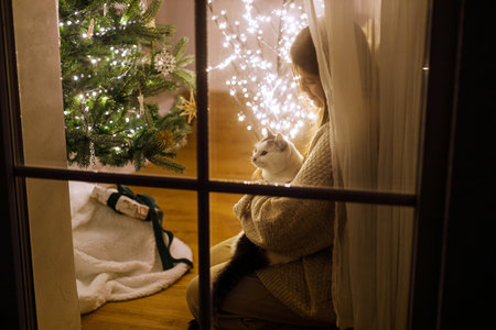 Beautiful woman in window hugging cute cat and looking at stylish gifts on background of festive living room with christmas tree lights. Atmospheric christmas eve at home. Merry Christmas!の写真素材