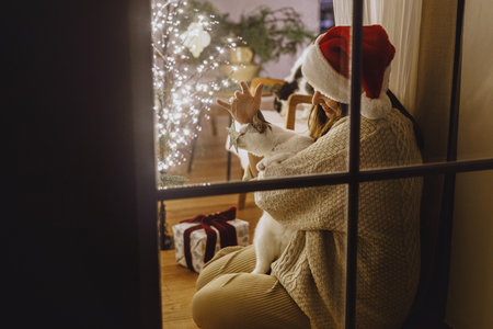 Merry Christmas! Happy woman in santa hat decorating stylish christmas tree and hugging cute cat in window on background of festive living room with lights. Atmospheric christmas eve at homeの写真素材