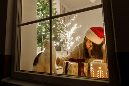 Merry Christmas! Happy woman in santa hat and cute cat in window with stylish gifts and glowing lantern on background of christmas tree with lights. Atmospheric christmas eve at home.の写真素材