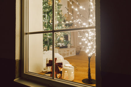 Merry Christmas! Stylish Christmas gift and glowing lantern in window on background of christmas tree with lights in evening. Atmospheric christmas eve at home. Holiday magical sceneの写真素材