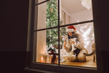 Merry Christmas! Happy woman in santa hat hugging cute cat in window on background of stylish gifts and christmas tree in festive living room with lights. Atmospheric christmas eve at homeの写真素材