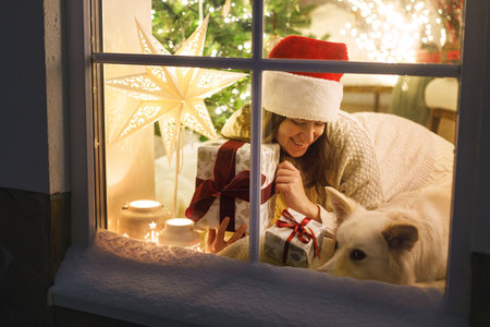 Merry Christmas! Happy woman in santa hat sitting with stylish christmas gift and cute dog in snowy window inside of festive room with christmas tree lights. Atmospheric christmas eve at homeの写真素材
