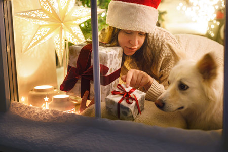 Merry Christmas! Happy woman in santa hat sitting with stylish christmas gift and cute dog in snowy window inside of festive room with christmas tree lights. Atmospheric christmas eve at homeの写真素材
