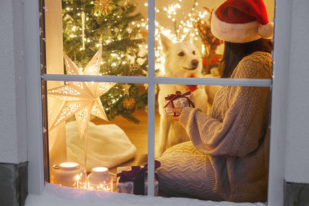 Merry Christmas! Happy woman in cozy sweater and santa hat sitting with cute dog in window in living room with christmas lights and gifts. Atmospheric christmas eve at home. Holiday magical sceneの写真素材