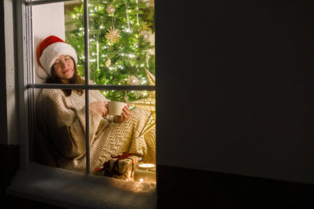 Happy woman in santa hat sitting with warm cup of tea in snowy window inside of festive room with christmas tree lights and gifts. Atmospheric christmas eve at home. Merry Christmas!の写真素材