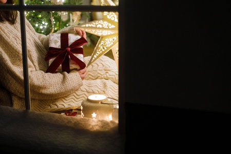 Happy woman holding stylish christmas gift in snowy window inside of festive room with christmas tree lights. Atmospheric christmas eve at home. Merry Christmas!の写真素材