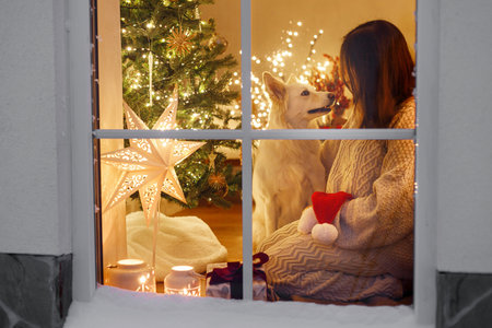 Happy woman in cozy sweater playing with cute dog and santa hat in window inside of festive living room with christmas tree lights and gifts. Atmospheric christmas eve at home. Merry Christmas!の写真素材