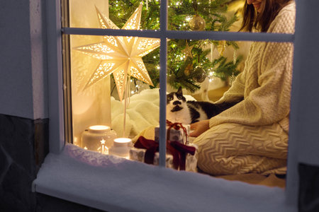 Merry Christmas! Happy woman in cozy sweater sitting with cute cat in snowy window on background of christmas lights and gifts. Atmospheric christmas eve at home. Holiday magical sceneの写真素材