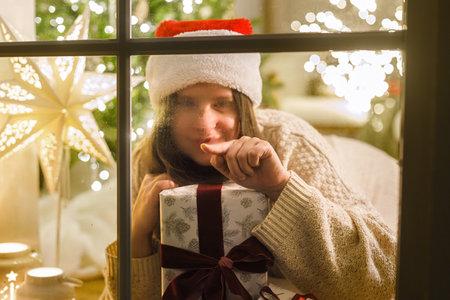 Merry Christmas! Happy woman in santa hat drawing heart with finger on window with stylish gift on background of festive lights. Atmospheric christmas eve at home. Holiday magical sceneの写真素材