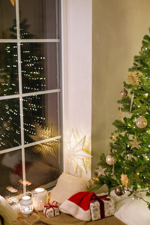 Stylish christmas gifts, glowing lantern, soft cozy pillow and santa hat under christmas tree with lights. Atmospheric christmas eve at home, winter hygge still life. Merry Christmas!の写真素材