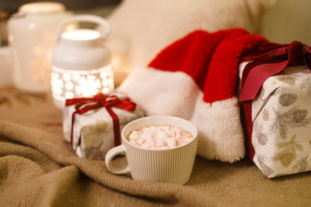 Winter hygge. Warm cup of cocoa with marshmallow under festive christmas tree with lights, gifts, soft cozy pillow and santa hat. Atmospheric christmas eve at homeの写真素材