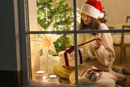 Happy woman in santa hat opening stylish christmas gift in snowy window inside of festive room with christmas tree lights. Atmospheric christmas eve at home. Merry Christmas!の写真素材
