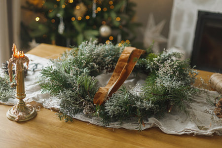 Making stylish Christmas rustic wreath. Wreath with evergreen branches, ribbon, scissors and candle on rustic table on background of atmospheric christmas tree. Winter holiday preparationsの写真素材