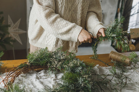 Making Christmas rustic wreath. Hands holding evergreen branches, making wreath on rustic table with pine cones, twine, scissors in atmospheric festive room. Winter holiday preparationsの写真素材