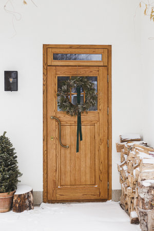 Stylish christmas rustic wreath with ribbon hanging on wooden doors, modern lantern, firewood and christmas tree at snowy entrance. Modern farmhouse outdoor decoration for winter holidayの写真素材