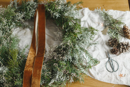 Making stylish Christmas rustic wreath. Wreath with fir and evergreen branches, ribbon, pine cones, scissors on rustic table in atmospheric festive room. Winter holiday preparationsの写真素材