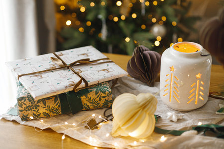 Stylish christmas glowing lantern, gift and modern paper ornaments on rustic table on background of christmas tree lights bokeh. Merry Christmas! Atmospheric christmas time, holiday bannerの写真素材