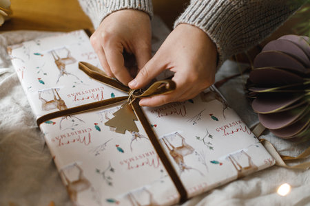 Woman wrapping stylish christmas gift on rustic table with modern paper ornaments and lights bokeh....