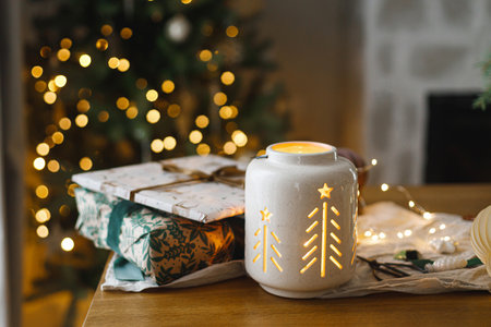Stylish christmas glowing lantern, gift and modern paper ornaments on rustic table on background of christmas tree lights bokeh. Merry Christmas! Atmospheric christmas time, holiday bannerの写真素材