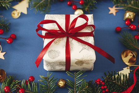 Merry Christmas! Stylish Christmas gift with red ribbon and green fir branches, red berries and gold ornaments on blue background. Xmas background. Season's greetingsの写真素材