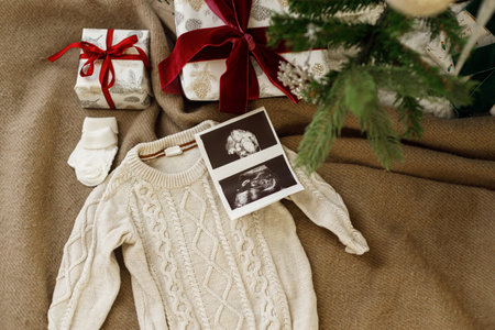 Pregnancy. Top view of stylish cute baby  knitted sweater, ultrasound scan and gift boxes under decorated christmas tree. Pregnant during christmas holidays. Fertility conceptの写真素材