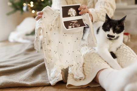 Happy pregnant woman holding ultrasound scan and cute baby clothes and sitting with cat at christmas festive decorated tree. Pregnancy during holidays, magical joyful time, best giftの写真素材