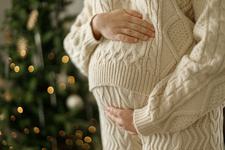 Happy pregnant woman in knitted outfit hugging belly close up on background of decorated christmas tree. Pregnancy during holidays, magical joyful timeの写真素材