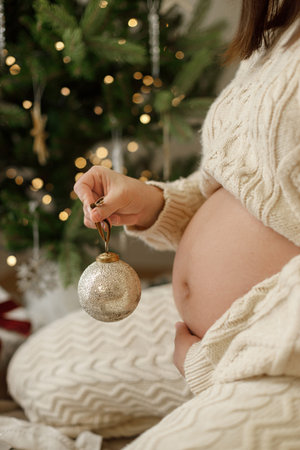 Stylish pregnant woman in knitted outfit hugging belly and holding festive bauble close up under...