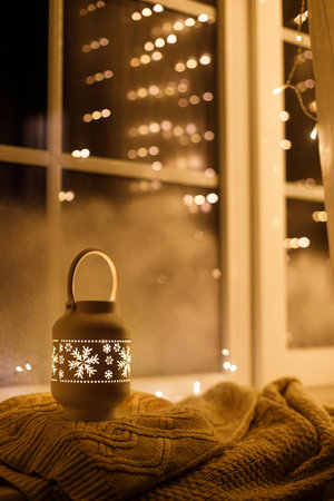 Merry Christmas! Stylish glowing christmas lantern and golden lights bokeh on cozy sweater at snowy window in evening. Atmospheric magical scene. Christmas holiday banner. Space for textの写真素材