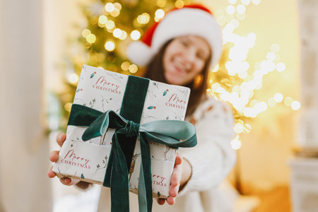Merry Christmas and Happy Holidays! Happy woman in santa hat holding christmas gift box on background of christmas tree with golden lights. Exchanging presents surpriseの写真素材