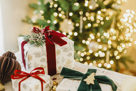 Merry Christmas and Happy Holidays. Stylish christmas gifts with spruce branch and modern ornaments on background of christmas tree golden lights. Christmas present holiday bannerの写真素材