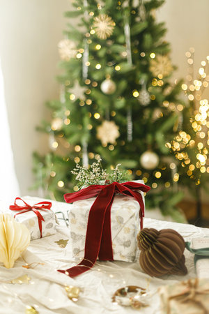 Merry Christmas and Happy Holidays. Stylish christmas gift box with red ribbon, spruce branch and modern ornaments on background of christmas tree lights. Christmas present holiday bannerの写真素材