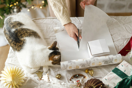Wrapping stylish christmas gift, winter holidays preparation. Hands in cozy sweater decorating gift box and and cute cat helping on table on background of decorated christmas tree with lightsの写真素材