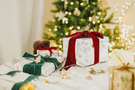 Merry Christmas and Happy Holidays. Stylish christmas gifts with spruce branch and modern ornaments on background of christmas tree golden lights. Christmas present holiday bannerの写真素材