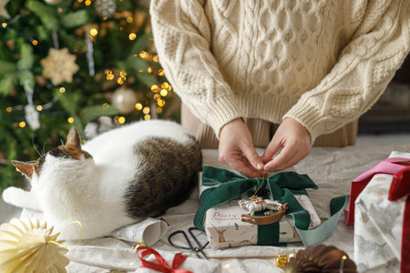 Wrapping stylish christmas gift, winter holidays preparation. Hands in cozy sweater decorating gift box and and cute cat helping on table on background of decorated christmas tree with lightsの写真素材